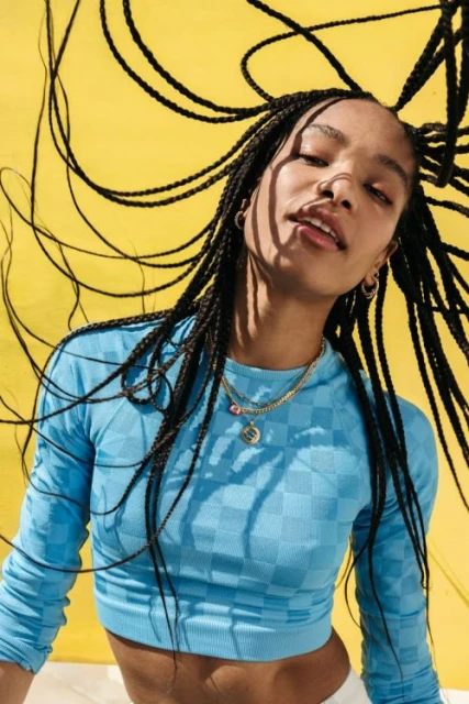 A young woman with braids poses in front of a yellow background, wearing a blue long-sleeved crop top and gold necklaces in a studio shot.