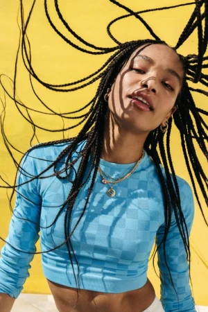 A young woman with braids poses in front of a yellow background, wearing a blue long-sleeved crop top and gold necklaces in a studio shot.