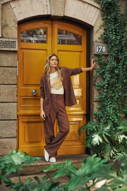 A stylish adult woman poses outdoors in a brown suit with a cream t-shirt, set against a yellow door and lush green foliage.