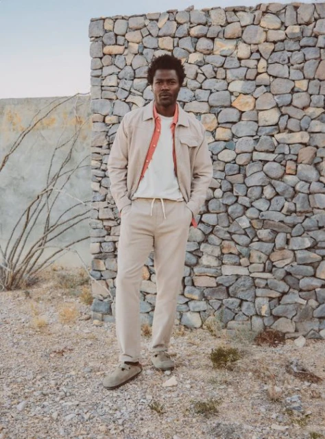 An African-American man poses outdoors in a tan jacket and pants, standing in front of a stone wall, in a fashion editorial shot.
