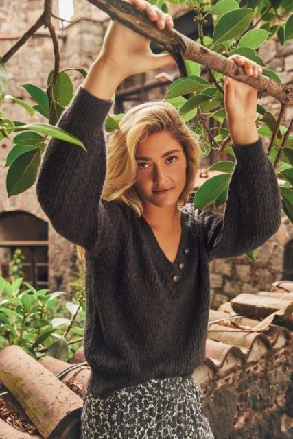 Young woman in a dark sweater poses outdoors near green foliage and a stone structure, looking at the camera with a neutral expression.