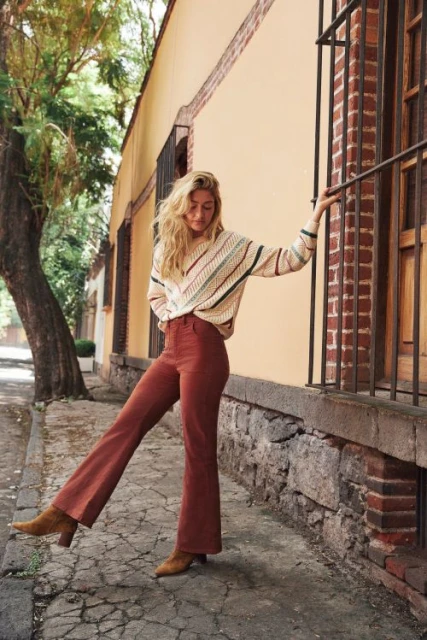 Fashionable woman in flared pants and a striped sweater stands on a sidewalk by a stucco building with green trees in the background.