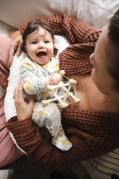 Smiling baby holding a wooden geometric toy while seated on a nursing pillow held by an adult in a brown waffle-knit cardigan.