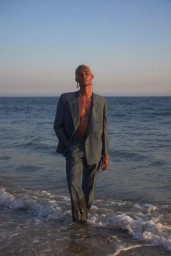 A man wearing a grey suit standing in ocean water at sunset, styled by Tennille Yamashita.