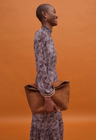 Model with shaved head wearing a high-neck patterned dress holding a large brown suede handbag against a warm orange backdrop