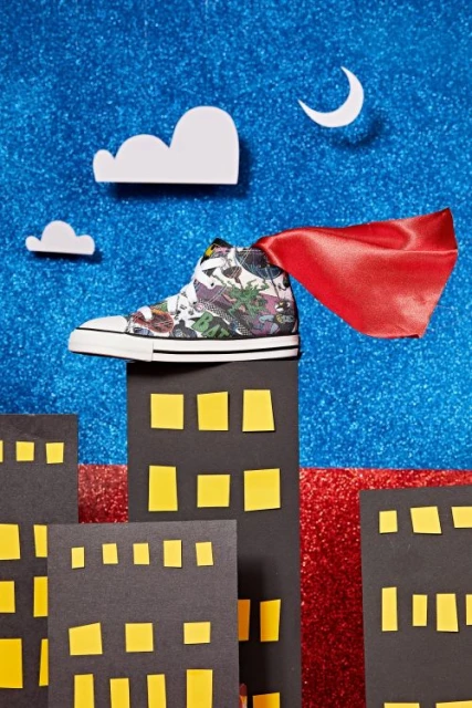 Sneaker with red cape on miniature cityscape against glittery backdrop. Fashion still life with creative styling and unique presentation.