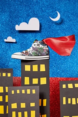 Sneaker with red cape on miniature cityscape against glittery backdrop. Fashion still life with creative styling and unique presentation.