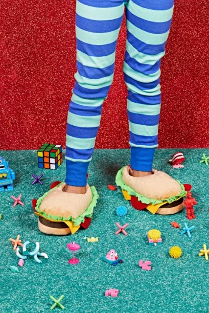Child's feet wearing burger slippers on a blue carpet with toys and a red glitter background, showcasing fun and playful footwear.