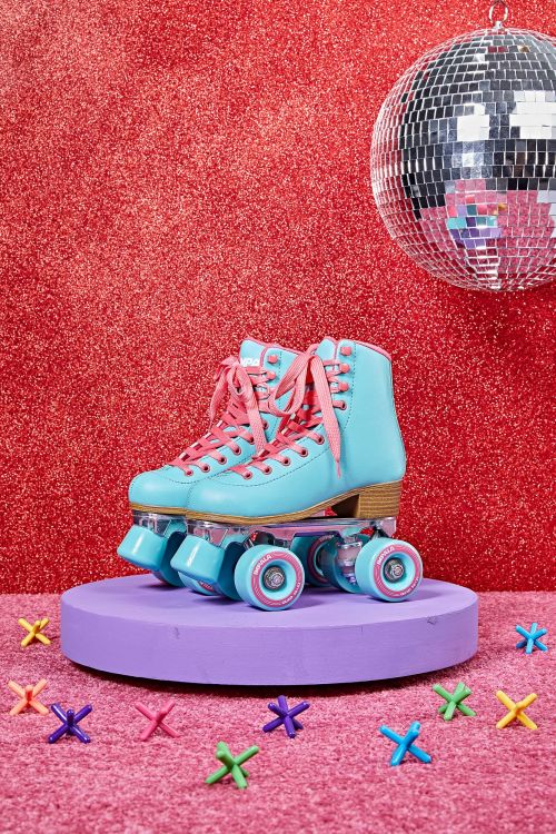 A pair of vintage turquoise roller skates are displayed on a purple platform against a red glitter backdrop with a disco ball.