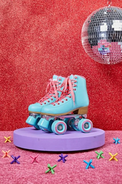 A pair of vintage turquoise roller skates are displayed on a purple platform against a red glitter backdrop with a disco ball.
