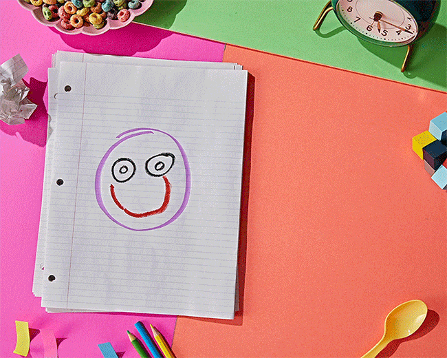 Top-down shot on colorful background: a lined paper stack with smiley face, desk clock, bowl of cereal, building blocks, writing utensils.