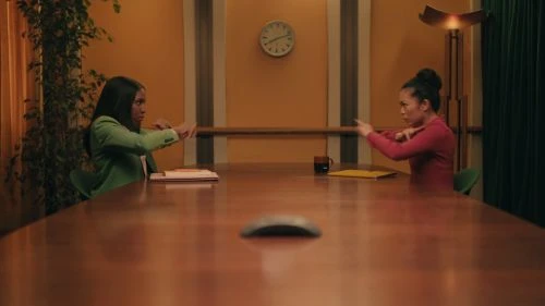 Two adult women mirror each other at a long wooden table in an office setting, wearing blazers and making gestures with their hands.