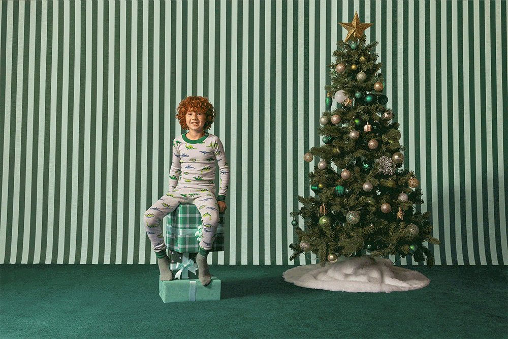 A child in pajamas sits on gift boxes beside a Christmas tree against a striped backdrop, creating a festive holiday scene.