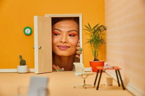 A woman with vibrant eyeshadow peers through a doorway in a room with peach and orange walls, a clock, and a small desk.
