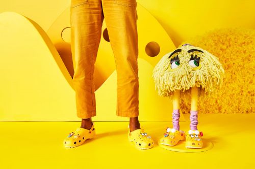 Person stands next to a yellow mop monster wearing Crocs shoes in a brightly lit, high-key yellow studio setting.