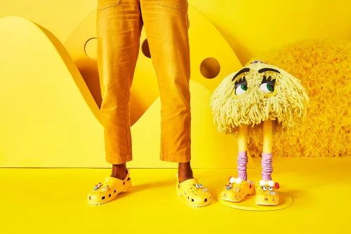 Person stands next to a yellow mop monster wearing Crocs shoes in a brightly lit, high-key yellow studio setting.
