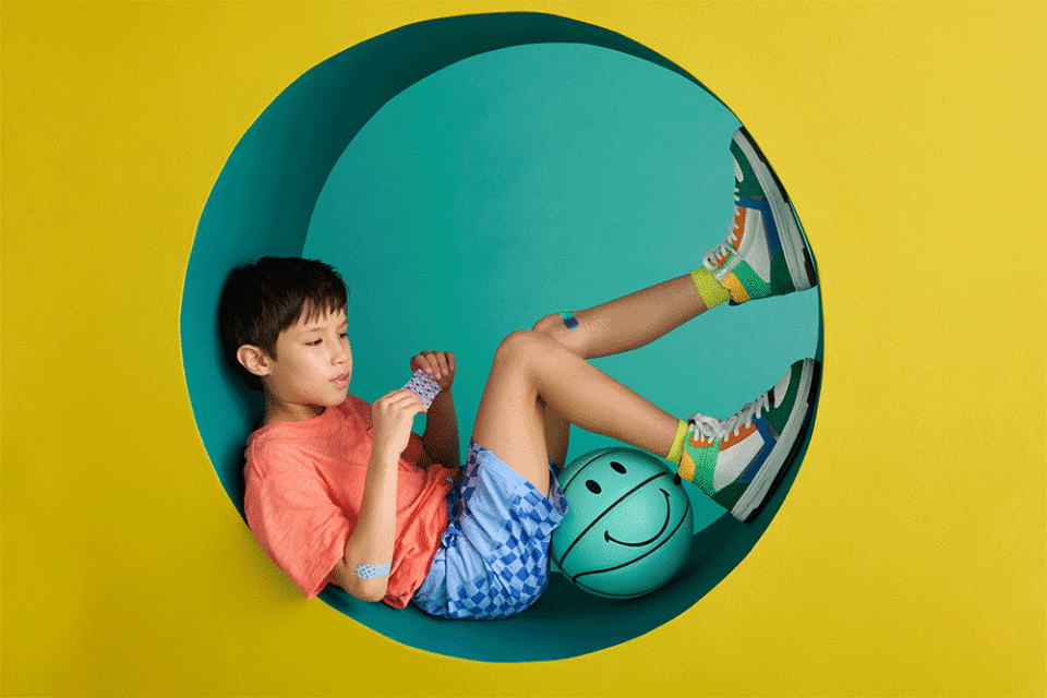 Young child sits in a teal circular cutout with a basketball and bandage in front of a yellow backdrop in a studio environment.