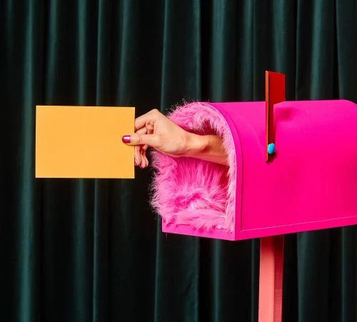 Hand places a yellow card into a pink mailbox with faux-fur trim, set against a dark green curtain backdrop in a studio setting.