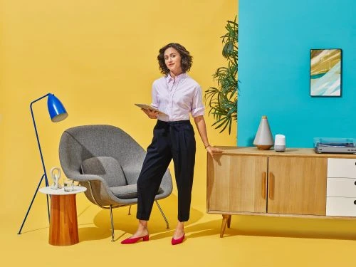 Stylish woman in pink shirt holds tablet in bright interior with blue floor lamp, gray chair, wooden cabinet, and vibrant yellow and blue walls.