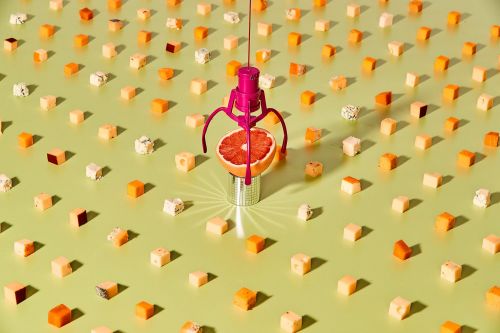 Elevated view of a grapefruit under a pink toy claw machine, surrounded by cheese cubes, creating a playful and colorful food still life.