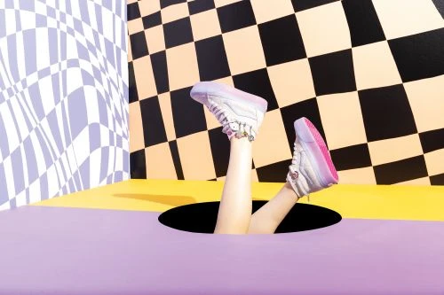 Person's legs and white sneakers sticking out of a hole in a floor with black, white, and yellow geometric background.