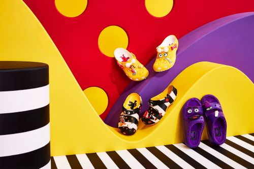 Colorful studio product shot of four pairs of decorated rubber sandals in yellow, purple, and black/white stripes against a bright backdrop.