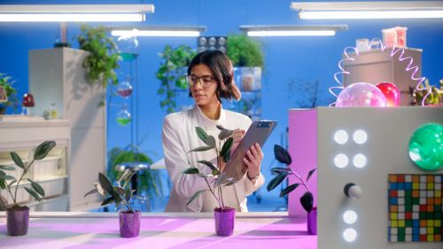 Woman in a white lab coat takes notes in a brightly lit laboratory filled with potted plants and scientific equipment.