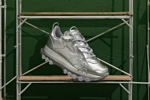 Silver Reebok sneaker on a wooden shelf against a green wall. The athletic shoe is metallic, offering a futuristic aesthetic.