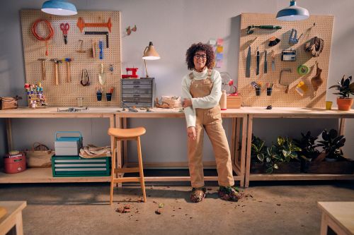 A person in overalls stands in a workshop with pegboards holding tools and workbenches with potted plants and toolboxes.