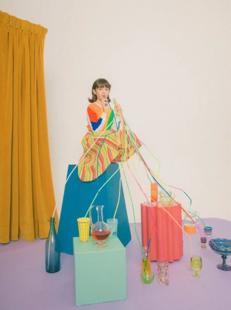 A young woman in colorful patterned clothing sits on a blue cube surrounded by drinks and neon tubing in a brightly lit indoor studio.