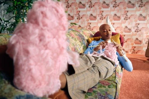 Man knitting pink yarn while sitting on floral couch in retro interior. Light-skinned man crafting pink fabric with needles.