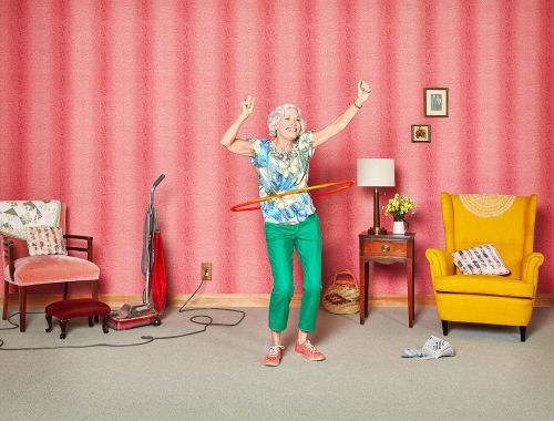 Smiling senior woman in a floral shirt and green pants playfully hula hooping in a room with pink wallpaper and vintage furniture.