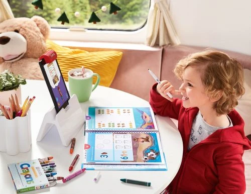 A young boy in a red hooded jacket is coloring in a workbook, sitting at a table with art supplies and a tablet device indoors.