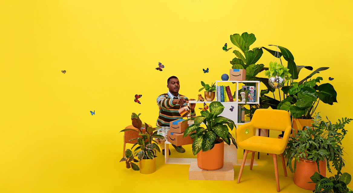 An African-American man sits amongst houseplants and Amazon boxes against a yellow background, sorting through a package with butterflies.