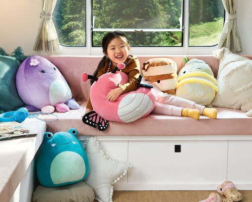 A happy Asian child sits on a pink sofa with a variety of plush pillows and cushions in an indoor setting with a window.