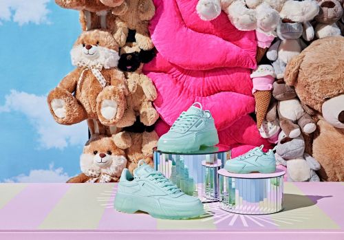Three light green Reebok sneakers displayed on iridescent platforms, surrounded by a backdrop of assorted plush toys.