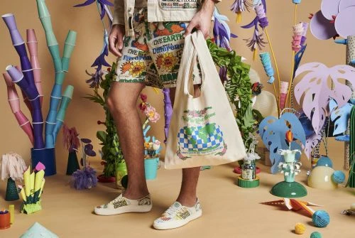 Man wearing patterned shorts and sneakers holding a tote bag surrounded by whimsical decorations against a beige background indoors.