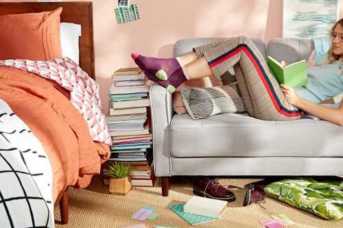 Woman reading a book while lying on a grey sofa in a casual indoor setting with books, shoes, and other items scattered around.