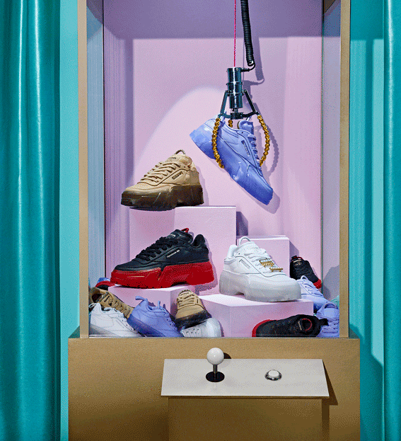 Eye-level shot of various sneakers inside a tan claw machine with turquoise curtains in the background, fashion-forward footwear.