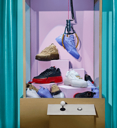 Eye-level shot of various sneakers inside a tan claw machine with turquoise curtains in the background, fashion-forward footwear.