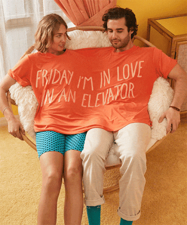 A smiling couple shares an oversized orange t-shirt with a funny slogan while sitting together in a wicker chair indoors.