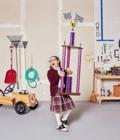 Young girl in school uniform excitedly holding a large trophy with checkered flags in a garage with yard tools and a wooden race car.