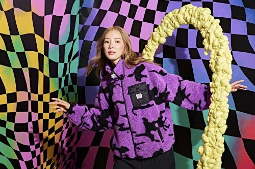 Woman posing in a purple camouflage fleece jacket against a vibrant checkered background with yellow foam structure in a studio setting.