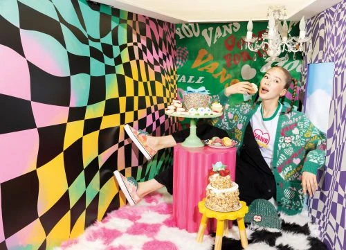 Woman in a whimsical room eating cake, wearing a green cardigan, next to cakes and a Vans beanie. The walls have a psychedelic pattern.