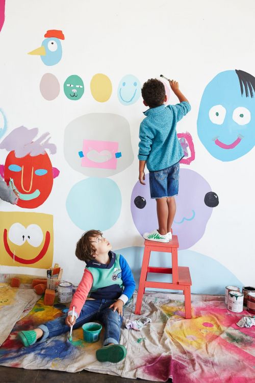 Two boys paint colorful faces and shapes on a white wall in an indoor setting, creating an expressive and playful art display with painting supplies.