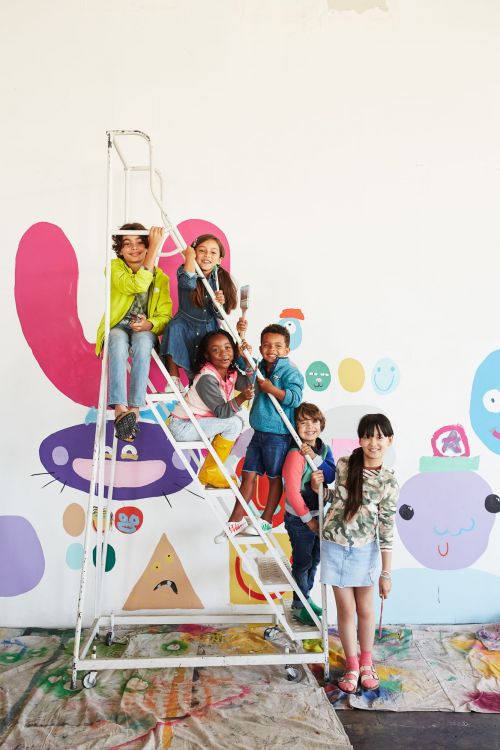 Group of smiling children on a rolling ladder with paintbrushes in a brightly lit art studio with colorful abstract mural on the wall.