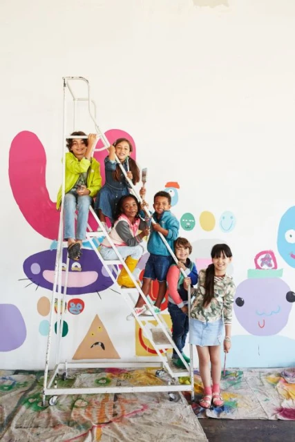 Group of smiling children on a rolling ladder with paintbrushes in a brightly lit art studio with colorful abstract mural on the wall.