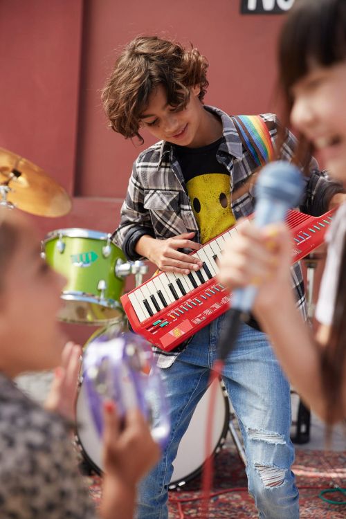 Young boy with an electric piano keyboard with musicians in the background playing the drums and microphone stand outdoors.