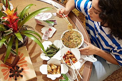Woman enjoying a takeout meal with noodles, dumplings, and stir-fry, sitting at a table with a woven placemat and a potted plant.