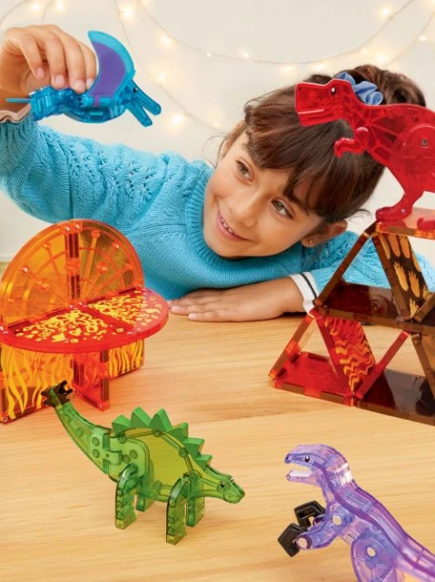 A child plays with translucent, colourful dinosaur figures on a table indoors near illuminated fairy lights in a lifestyle setting.